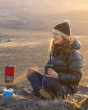 An adult smiling and enjoying a hot drink in their Klean Kanteen 12oz Insulated Camping Mug, sat outside in the sunshine on a mountain