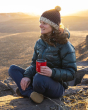An adult is sitting on a rock, on top of a hillside with the sun setting in the background, whilst holding their Klean Kanteen Camping Mug in Red