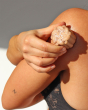 A person rubbing the Lamazuna Organic Sublime Orange Blossom Body Shimmer Solid Bar onto the skin on the shoulder and back of the arm, showing the natural shimmer in the sunlight