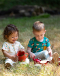 Two children playing happily together with a red tea set in a grassy field. The child on the right is wearing the Little Green Radicals Garden Birds Organic T-Shirt & Jogger Playset, made from GOTS Organic Cotton, this cosy jogger set come with light blue