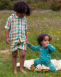 Two children together in a grassy field with yellow flowers in the background. The child on the right is wearing the Little Green Radicals Garden Birds Organic Cotton Zip Babygrow, and the child on the left is wearing the Little Green Radicals Rainbow Str