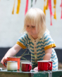 A child playing with a red set of cups outside, wearing the Little Green Radicals Sail Away Knitted Shortie. Made from GOTS Organic Cotton, this shortie has a beautiful blue and cream wavy stripe with yellow arm and leg cuffs, with button on the chest and