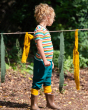 A child stood, in front of a rope line with colourful cloths tied onto it and looking into the distance, wearing the Little Green Radicals Rainbow Stripes Summer Short Sleeve T-Shirt.