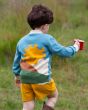 A child is standing in a grassy field and is holding a red cup whilst wearing the Little Green Radicals Organic Cotton From One To Another Sunshine Knitted Cardigan, which shows the sunshine design on the back
