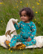 A child sat in a grassy field on a fluffy blanket and playing with soft plush toys, is wearing the Little Green Radicals Garden Birds Organic Cotton Zip Babygrow, made from GOTS Organic Cotton, this cosy a teal footed babygrow has fun garden birds design,