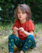 A child sitting down on a small grassy hill, wearing the Little Green Radicals Organic Cotton Little Lizard Comfy Joggers. Made with GOTS Organic Cotton, these joggers are a lovely deep forest green with a playful chameleon lizard print, a drawstring cord