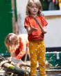Two children playing outside near a log pile, The child in front is playing with a stick and is wearing an orange top and Little Green Radicals Counting Sheep Organic Comfy Joggers. Made from GOTS Organic Cotton, they comfy joggers are orange with sheep p