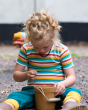 A child sitting down on the ground playing with small pebbles in a yellow bucket, wearing the Little Green Radicals Rainbow Stripes Summer Short Sleeve T-Shirt. Made from GOTS Organic Cotton, this fun striped t-shirt comes with white, green, yellow, red a