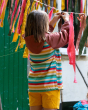 The back of the Little Green Radicals Rainbow Striped Recycled Waterproof Windbreaker Jacket, worn by a child playing with coloured fabric outside