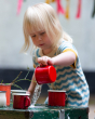 A child pouring water from a red cup onto a painted table, wearing the Little Green Radicals Sail Away Knitted Shortie. Made from GOTS Organic Cotton, this shortie has a beautiful blue and cream wavy stripe with yellow arm and leg cuffs, with button on th