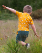 A child happily playing in a grassy field with yellow flowers, wearing the Little Green Radicals Counting Sheep Short Sleeve T-Shirt. Made from GOTS organic cotton, this t-shirt has a fun sheep print on orange fabric