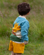 A child is standing in a grassy field and is wearing the Little Green Radicals Organic Cotton From One To Another Sunshine Knitted Cardigan, whilst showing the sunshine design on the back