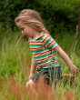A child walking in a long, grassy field wearing the Little Green Radicals Rainbow Stripes Summer Short Sleeve T-Shirt, showing the poppers open on the shoulder