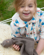 A child happily playing and holding the Little Green Radicals Bear Organic Cotton Soft Toy, on a fluffy cream rug and grassy background
