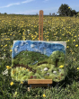A colourful outdoor background filled with bright yellow flowers, and The Makerss - Warm Landscape Needle Felt Crafting Kit set up on an easel.