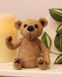 The Makerss Needle Felt Jointed Bear. A beautifully crafted pose-able light brown bear, with a brown nose and black stick in eyes, sat on a  shelf in front of a candle and green plant