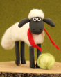 A close of of a crafted Shaun The Sheep On Four Legs Needle Felt character with a fluffy felted body, red scarf and green lettuce, on a green background and stood on a wooden base