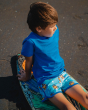 A child sitting on a surf board on the sand, whilst wearing a Solid Blue Maxomorra T-shirt and Maxomorra Children's Organic Cotton Monkey Runner Shorts