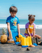 Two children building sandcastles on a sunny beach. The child on the left is wearing the Maxomorra Monkey Raglan top and blue shorts, and the child on the right is wearing the Maxomorra Children's Organic Cotton Pineapple Raglan Short Sleeve Top and purpl