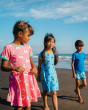 Three children taking a walk on the beach. The child on the left is wearing the Maxomorra Unicorn Organic Cotton Children's Short Sleeve Circle Dress, the child in the middle is wearing a Maxomorra Dolphin Dress, and the child on the right is wearing a na