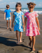 Three children walking on the beach. The child in front is wearing the Maxomorra Unicorn Organic Cotton Children's Short Sleeve Circle Dress, the child in the middle is wearing a Maxomorra Dolphin Dress, and the child at the back is wearing a navy blue t-
