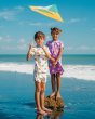Two children flying a kite at the beach on a sunny day. The child in front is wearing the Maxomorra Children's Organic Cotton Turtle Pyjama Set, and the child behind is wearing a Maxomorra lemon dress