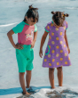 Two children gazing at the waves under their feet. The child on the left is wearing the Maxomorra Children's Organic Cotton Unicorn Raglan Short Sleeve Top and green Maxomorra muslin shorts, and the child on the right is wearing a Maxomorra lemon dress