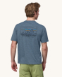An adult wearing the Patagonia Men's Capilene Cool Daily Graphic Shirt - 73 Skyline / Utility Blue X-Dye, with a Patagonia mountain range logo and "Save Our Home Plantet" text underneath, on the back of the t-shirt. Adult is also wearing light green short