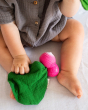 Close up of the Oli & Carol eco-friendly mini radish baby comforter toy on a grey sheet next to a baby's leg
