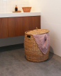 Olli Ella natural woven rattan laundry basket on a grey bathroom floor in front of some cupboards