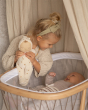 A child leans over a baby siblings cot holding the Olli Ella Lullaby Dozy Dinkum Doll Lyra.