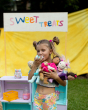 A child happily eating cake whilst holding the Dinkum Sweet Treat collection in their arms. In the background is a yellow sheet, colourful box shelves and a "Sweet Treats" sign