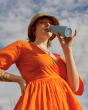 An adult in a bright, orange dress taking a sip from their One Green Bottle 350ml Evolution Collection Bottle - Sports Cap in Ocean, with a blue sky background