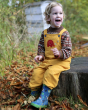 A child happily sat on a tree stump outside wearing the Yellow Frugi x Babipur natural organic cotton cord dungarees with red elephant