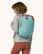 An adult carrying the Patagonia Atom Tote Backpack 20L - Skiff Blue on their back, showing the bag shoulder straps, bag carry handles, zipper and side pocket, on a cream background