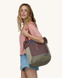 An adult carrying the  Patagonia Ultralight Black Hole Tote Pack on their shoulder. Pictured is purple and light/dark beige bag, with salmon pink carry straps and a royal blue zipper