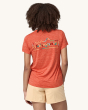 An adult showing the back of the the Patagonia Women's Capilene Cool Daily Graphic Shirt - Unity Fitz / Pimento Red X-Dye, with cream shorts on a cream background. The back of the t-shirt has the Patagonia logo in differnt colours consisting of yellow, re