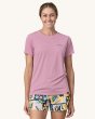 An adult wearing the Patagonia Women's Capilene Cool Daily Graphic Shirt - Boardshort Logo / Milkweed Mauve X-Dye, showing black Patagonia writing on the front of the t-shirt, and colourful tropical shorts