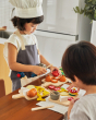 Young girl dressed as a chef playing with the PlanToys wooden play food cheese and charcuterie board set on a wooden worktop 