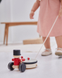 Close up of young girl in a pink dress pulling a PlanToys musical pull along drummer toy on a white floor