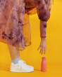 A person in a floaty, pink and purple tie dye dress, reaching down to pick up their One Green Bottle 350ml Evolution Collection Bottle - Sports Cap in Coral, on a bright yellow background
