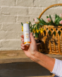 A person holding the Green People Natural and Organic Mineral Suncream SPF30 Scent Free bottle in their hand, with flowers in a basket, in the background