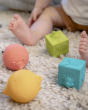 The rubber balls and cubes of the Sophie The Giraffe - So Pure Early Learning Gift Set, laid on a cream fluffy rug with a child sat in the background