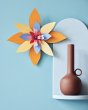 Studio Roof orange and blue craft flower hanging on a blue wall next to a light blue shelf with a brown candle holder