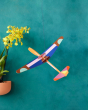 The Studio ROOF Giant Glider Plane, a cardboard model plane with geometric patterns and bright colours, put together, hanging with a mottled teal background, an orange plant pot containing green plant with bright yellow flowers is to the left. 
