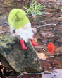 The Makerss Needle Felt Fishing Gnome. A beautifully crafted fishing gnome with a green hat, white bushy beard, little red wellington boots, a small fishing stick and a small red fish, sat on a rock at the riverside