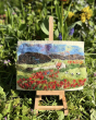 A bright, grassy green background with yellow and purple flowers, with the Makerss Poppy Landscape Needle Felt Kit on an easel in the forground