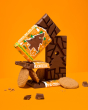 3 Tony's milk chocolate gingerbread bars stacked in a pile with some gingerbread biscuits on an orange background