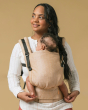 Woman stood on a beige background with a baby in a Tula forward facing baby carrier