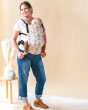 Woman stood in front of a beige wall carrying a baby in a Tula charmed free to grow baby carrier